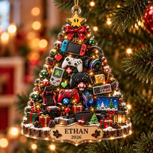 May include: A triangular Christmas tree ornament featuring miniature gaming consoles, controllers, a gaming chair, and headsets. The base is a wooden plaque with the name ETHAN and the year 2026 engraved, decorated with holly leaves and berries.