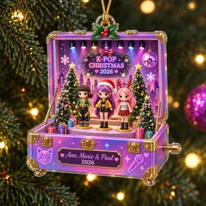 Könnte beinhalten: Ein K-Pop Weihnachtsornament 2026 in Form eines offenen violetten Koffers mit goldenen Akzenten. Im Inneren stehen drei Chibi-Puppen auf einer Bühne zwischen zwei geschmückten Weihnachtsbäumen. Vorne steht der Text Ann Marie & Paul 2026.