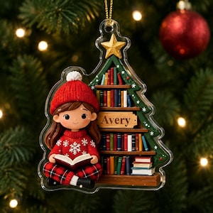 May include: A clear acrylic Christmas tree ornament featuring a brown-haired character in a red beanie and snowflake sweater reading a book. Beside the character is a bookshelf with the name Avery printed on a wooden plaque and a gold star on top.