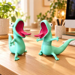 May include: Two light green lizard-shaped figures with wide-open mouths, bright pink interiors, and extended pink tongues. Each figure has large white eyes with black pupils, small arms, and a long tail. One figure features a ribbed midsection.