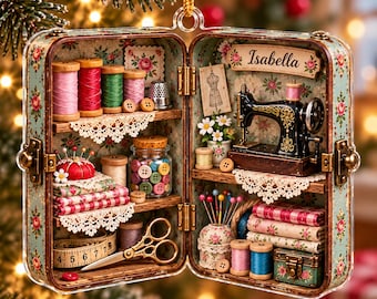 May include: A miniature sewing room inside an open suitcase ornament featuring the name Isabella, a black sewing machine, colorful thread spools, fabric stacks, a pincushion, buttons in a glass jar, scissors, and a measuring tape.