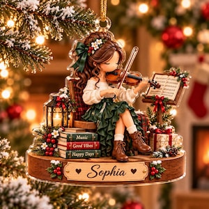 Könnte beinhalten: Ein dekoratives Acryl-Ornament, das ein Mädchen beim Geigenspiel zeigt, sitzend auf einem Stuhl mit Büchern, beschriftet mit Music, Good Vibes und Happy Days. Der Holzsockel trägt den eingravierten Namen Sophia und ist mit Stechpalmen verziert.