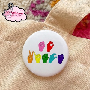 May include: A white button pin with the words "VOTE" spelled out in colorful American Sign Language hand gestures. The pin is set against a beige fabric background. The pin has a glossy finish.