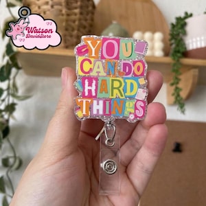 May include: A colorful badge reel with the text "YOU CAN DO HARD THINGS" in various bright colors. The badge reel has a clear plastic holder and a silver-colored metal clip. The badge reel is decorated with glitter.