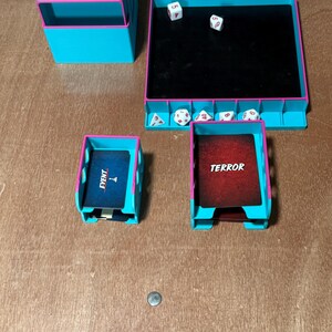May include: A tabletop game set with teal and pink accents. Includes a dice rolling tray, card holders with "EVENT" and "TERROR" cards, dice, and a storage box. The set is made of plastic.