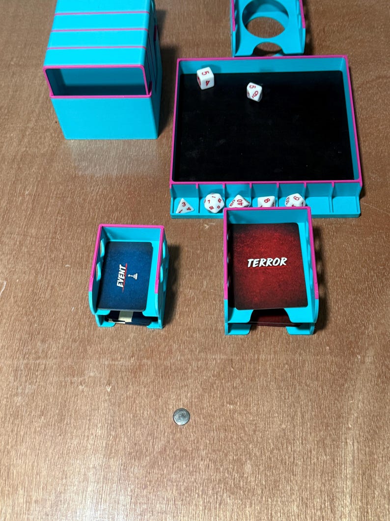 May include: A tabletop game set with teal and pink accents. Includes a dice rolling tray, card holders with "EVENT" and "TERROR" cards, dice, and a storage box. The set is made of plastic.