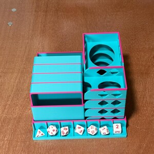 May include: A teal and pink dice tower and storage unit. The unit has multiple compartments, including a dice tray, stacked storage, and a tower with a circular opening. Several white dice with black numbers are in the tray.