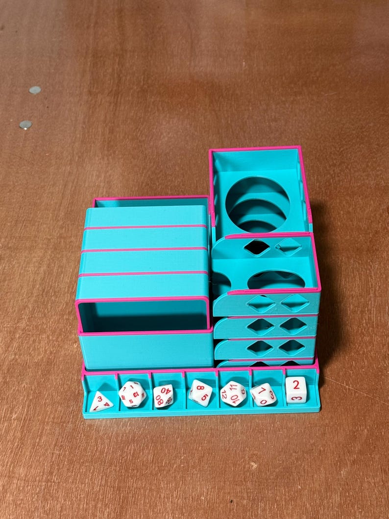 May include: A teal and pink dice tower and storage unit. The unit has multiple compartments, including a dice tray, stacked storage, and a tower with a circular opening. Several white dice with black numbers are in the tray.