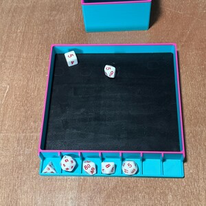 May include: A teal and pink dice rolling tray with a black interior, and a matching storage container. Several white dice with red numbers are visible inside and next to the tray. The dice are used for tabletop games.