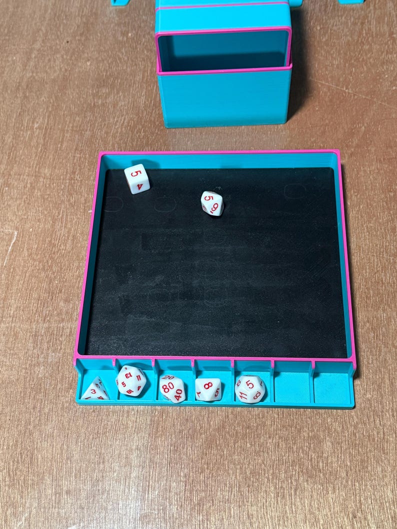 May include: A teal and pink dice rolling tray with a black interior, and a matching storage container. Several white dice with red numbers are visible inside and next to the tray. The dice are used for tabletop games.