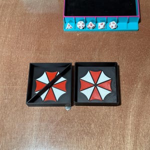 May include: Two black square trays with the Umbrella Corporation logo in red and white. One tray has a diagonal line across the logo. A dice tray with dice and a coin are also visible.