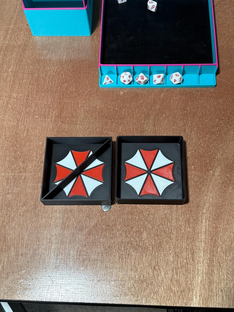 May include: Two black square trays with the Umbrella Corporation logo in red and white. One tray has a diagonal line across the logo. A dice tray with dice and a coin are also visible.