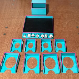 May include: A collection of teal and pink 3D-printed tabletop gaming accessories. Includes a dice tray, dice box, and individual dice holders. The dice tray has a black interior. Dice are visible inside the tray. The accessories are arranged on a wooden surface.