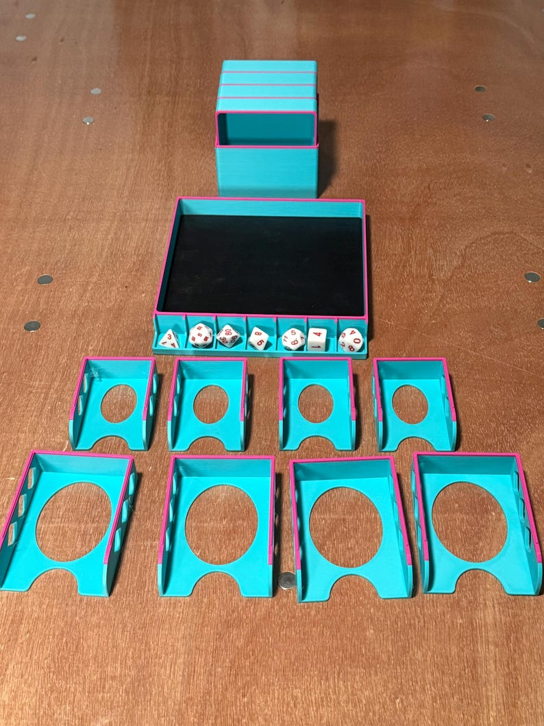 May include: A collection of teal and pink 3D-printed tabletop gaming accessories. Includes a dice tray, dice box, and individual dice holders. The dice tray has a black interior. Dice are visible inside the tray. The accessories are arranged on a wooden surface.