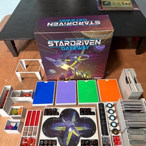 Puede incluir: Una caja de juego de mesa Stardriven Gateway con imágenes espaciales y texto, acompañada de varios componentes de juego que incluyen organizadores de plástico blanco, cajas de cartas de colores, pequeñas naves espaciales de plástico, dados negros y rojos.