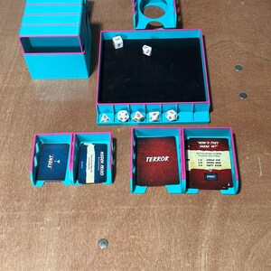 May include: A teal and pink tabletop game organizer with dice, cards, and storage compartments. The organizer includes dice trays, card holders, and a central play area. Cards are labeled "EVENT", "HIDDEN PRIZES", "TERROR", and "HOW'D THEY SNEAK IN?"