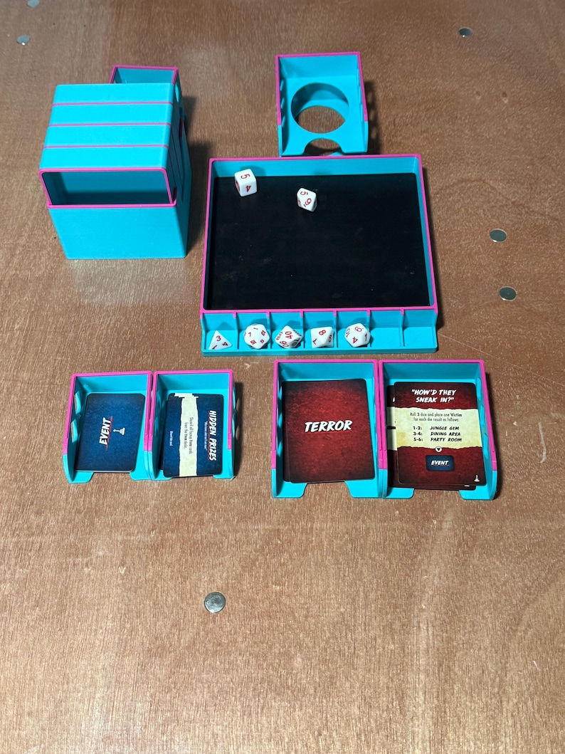 May include: A teal and pink tabletop game organizer with dice, cards, and storage compartments. The organizer includes dice trays, card holders, and a central play area. Cards are labeled "EVENT", "HIDDEN PRIZES", "TERROR", and "HOW'D THEY SNEAK IN?"