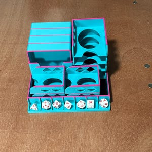 May include: A teal and pink dice tower and storage unit. The unit has multiple compartments and slots for dice. Several white dice with black numbers are in the tray. The unit is made of plastic.