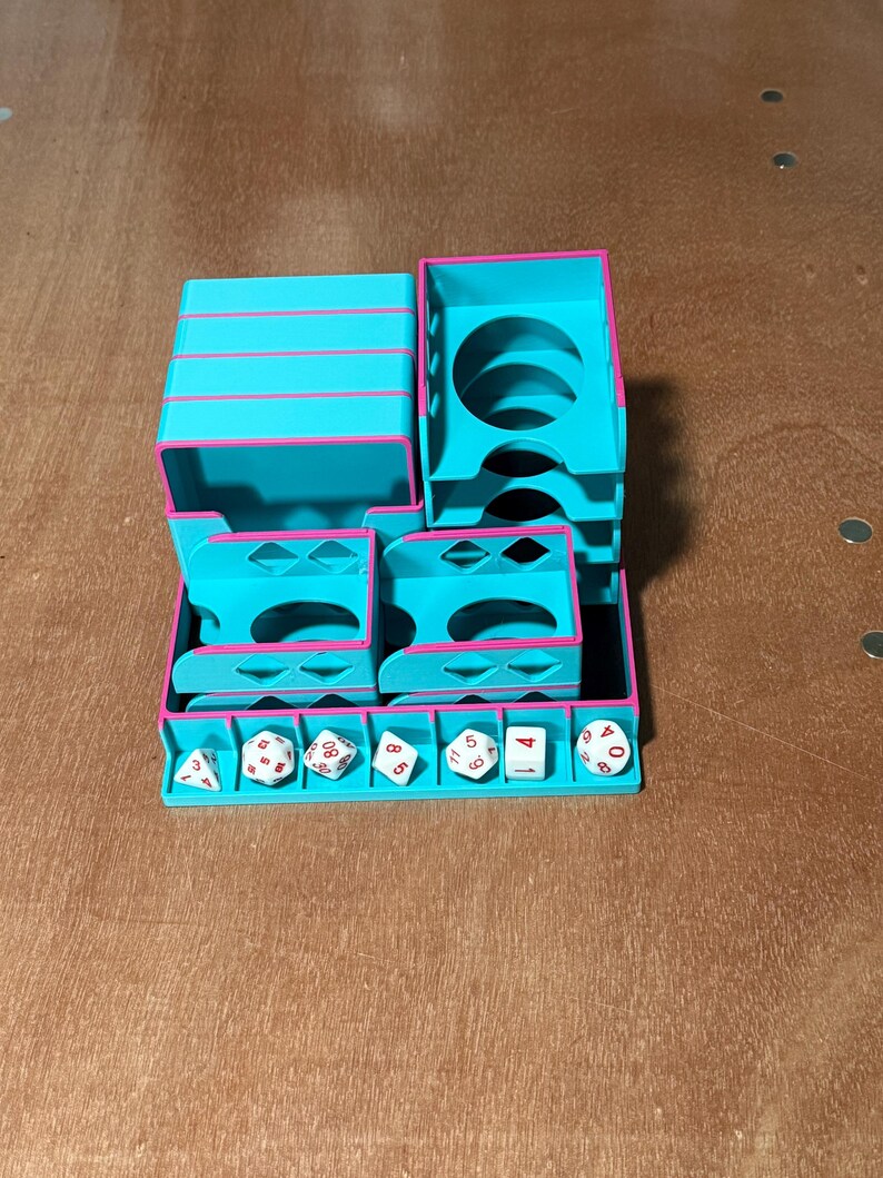 May include: A teal and pink dice tower and storage unit. The unit has multiple compartments and slots for dice. Several white dice with black numbers are in the tray. The unit is made of plastic.