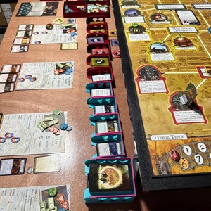 May include: A board game setup on a wooden table. The game includes a large game board, cards, tokens, and various storage containers in red, blue, and teal. The game board has a map design with text and illustrations.