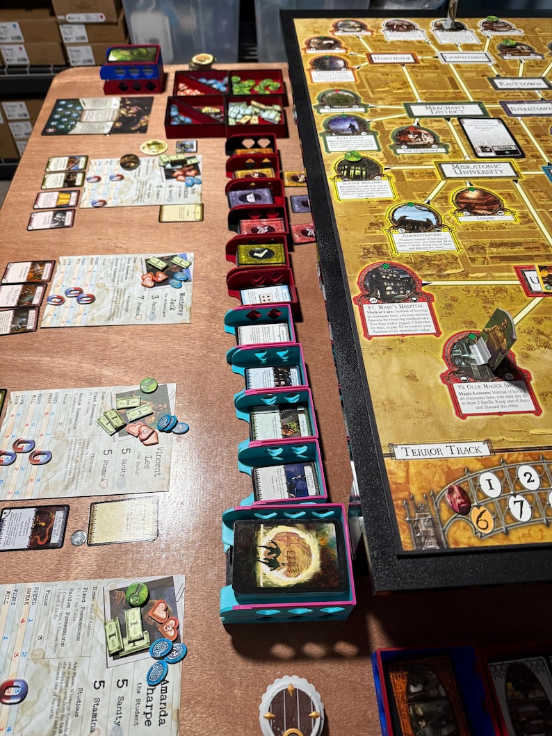 May include: A board game setup on a wooden table. The game includes a large game board, cards, tokens, and various storage containers in red, blue, and teal. The game board has a map design with text and illustrations.