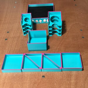 May include: A teal and pink tabletop gaming organizer set. The set includes a tray with a black interior, two dice towers, a small box, and four trays. Dice are visible in the tray. The organizer is made of plastic.