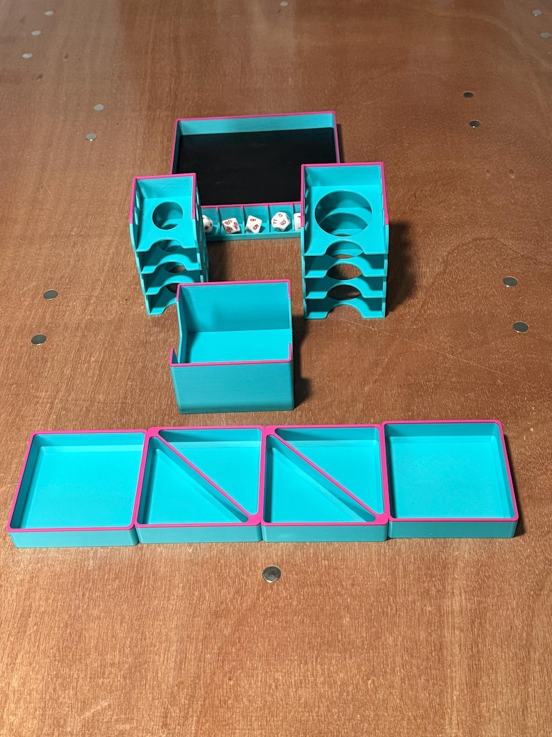 May include: A teal and pink tabletop gaming organizer set. The set includes a tray with a black interior, two dice towers, a small box, and four trays. Dice are visible in the tray. The organizer is made of plastic.