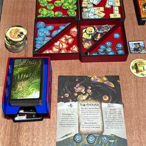 May include: A board game setup featuring red and black trays filled with game pieces, including tokens, cards, and money. A blue card holder is on the left, and a game card with text is in the center. The game pieces are in various shapes and colors.