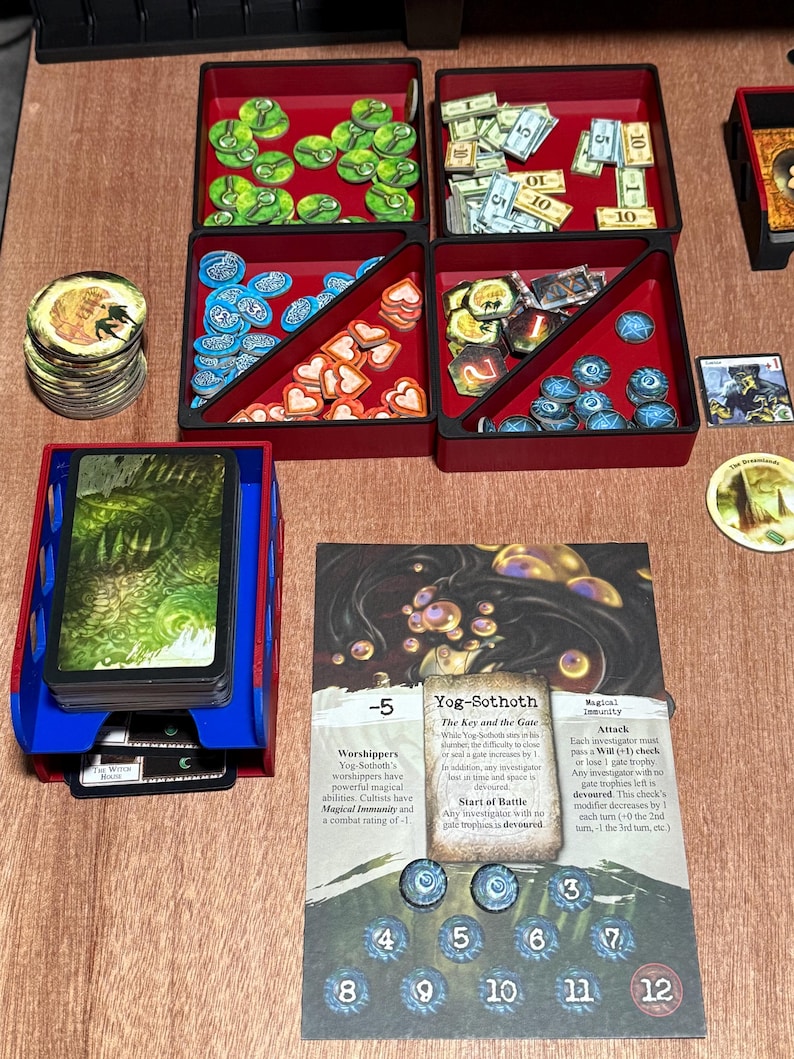 May include: A board game setup featuring red and black trays filled with game pieces, including tokens, cards, and money. A blue card holder is on the left, and a game card with text is in the center. The game pieces are in various shapes and colors.