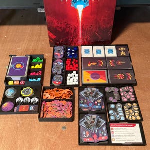 May include: A board game titled Revenant featuring a large game box with a red and black illustration of a spaceship, accompanied by multiple black plastic organisers filled with various game components including cards, tokens, and small coloured cubes.