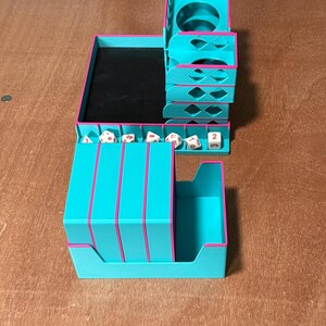 May include: A teal and pink tabletop organizer for dice and other gaming accessories. The organizer has multiple compartments, including a dice tray, a storage area, and a cup holder. The edges are trimmed in pink.