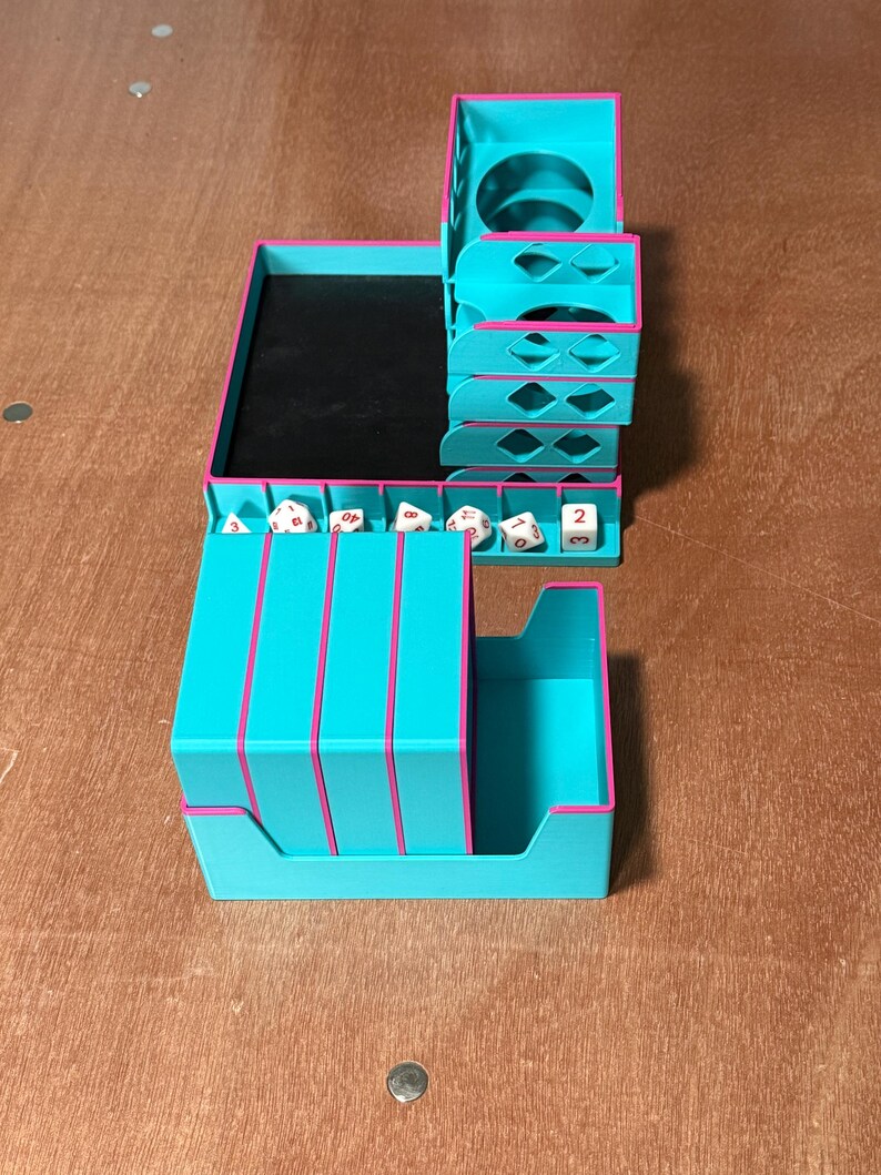 May include: A teal and pink tabletop organizer for dice and other gaming accessories. The organizer has multiple compartments, including a dice tray, a storage area, and a cup holder. The edges are trimmed in pink.