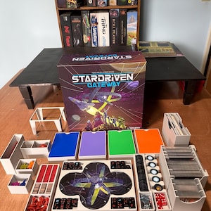以下が含まれることがあります： ボードゲーム「Stardriven Gateway」の箱が、白いプラスチックトレイ、赤いミニチュアフィギュア、黒と白のサイコロ、カードの束、宇宙をテーマにしたグラフィックが描かれた円形のゲームボードなど、整理されたゲームコンポーネントの背後に配置されています。
