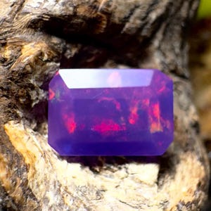 Puede incluir: Una gema rectangular facetada con un tono púrpura intenso y destellos internos rojos y anaranjados. La gema se encuentra sobre un fondo de madera natural texturizada, resaltando sus colores vibrantes y claridad.