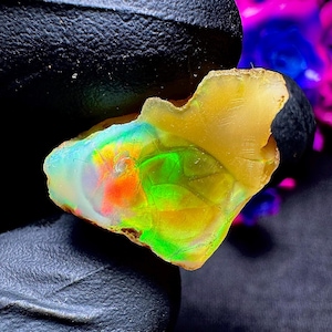 Puede incluir: Un ópalo en bruto y translúcido que presenta un vibrante juego de colores con destellos de verde neón, naranja brillante y amarillo sobre una base pálida semitransparente. La piedra tiene una forma irregular y dentada con texturas visibles.
