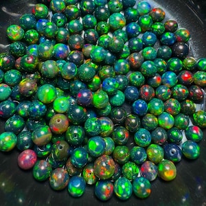 May include: A collection of round, dark-toned beads featuring a vibrant play of colour with iridescent flashes of green, red, and blue. Each bead has a central hole, and the surface displays a polished, reflective finish with varied patterns.