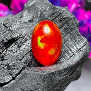 May include: An oval gemstone, predominantly red with yellow and green inclusions. The polished stone is displayed on a dark, textured surface. The gemstone's vivid colours are accentuated by the dark backdrop. Suitable for jewellery.