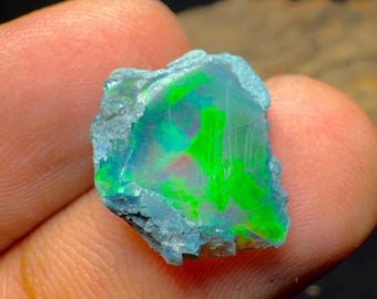 May include: A rough-cut opal gemstone with vibrant internal colors. The stone displays flashes of green, blue, and red within a translucent matrix. The outer edges are a muted blue-gray, contrasting with the bright interior.