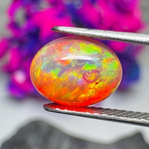 May include: An oval-shaped cabochon opal gemstone displaying a vibrant play-of-color with dominant flashes of neon orange, bright yellow, and electric green, held securely between the serrated metal tips of a pair of silver-toned tweezers.