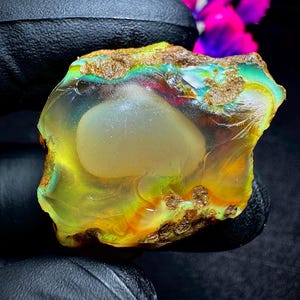 May include: A raw, translucent opal gemstone featuring a central milky white inclusion surrounded by vibrant flashes of neon green, yellow, and orange play-of-color, with rough, earthy brown matrix edges along the top and bottom borders.