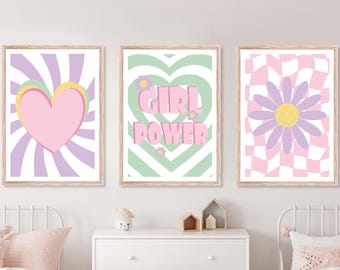 May include: Three framed art prints with a retro theme. The prints feature a pink heart, the words "GIRL POWER", and a daisy with a smiley face. The color palette includes pink, purple, yellow, and green. The prints are displayed above a white dresser.