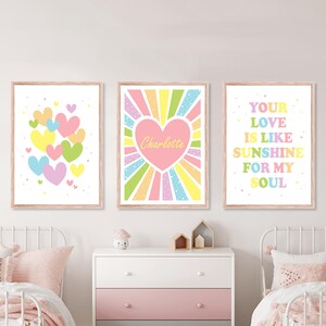 May include: Three framed wall art prints with light wood frames. The left print features colourful pastel hearts. The centre print displays a large pink heart with the name Charlotte in yellow. The right print reads Your Love Is Like Sunshine For My Soul.