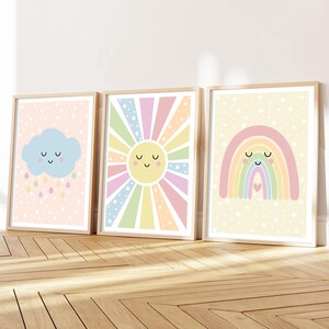 May include: Three framed wall art prints featuring pastel illustrations. The first shows a blue cloud with raindrops on a pink dotted background. The second depicts a yellow sun with radiating colourful stripes. The third shows a rainbow with a heart.