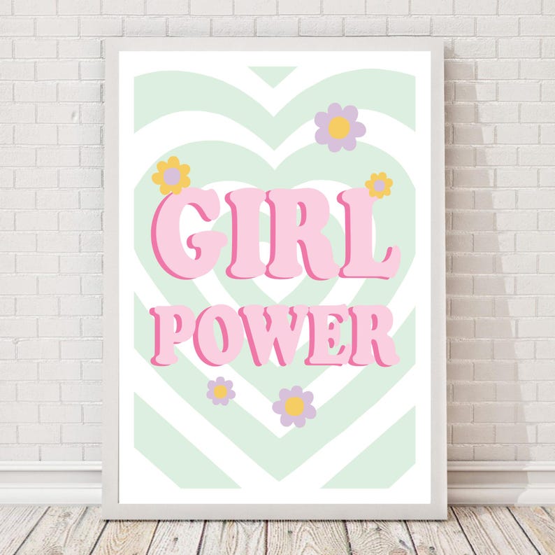 May include: A framed print with the words "GIRL POWER" in pink, set against a light green and white heart-shaped background. The design includes small flower illustrations in yellow, purple, and white.