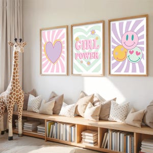 May include: Three framed wall art prints with a retro theme. The prints feature a heart, the words "GIRL POWER", and smiley faces. The color palette includes pink, purple, yellow, and green. The prints are in wooden frames.