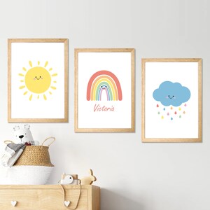 May include: Three framed wall art prints featuring a yellow sun with a face, a colourful rainbow with the name Victoria written below it, and a blue cloud with multicoloured raindrops. Each print is enclosed in a light wood frame.