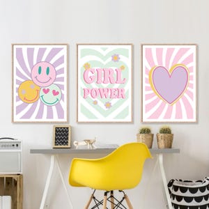 May include: Three framed wall art prints with a retro theme. One print features smiley faces, another reads "GIRL POWER," and the third has a heart design. The prints are in pastel colors with a wooden frame.