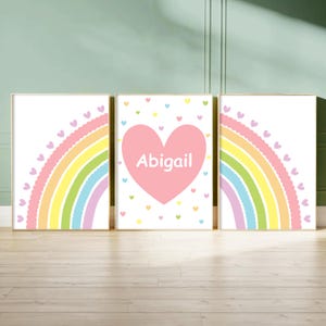 May include: A set of three framed wall art prints. The two outer prints feature a rainbow with scalloped edges and small heart accents. The centre print displays a large pink heart with the name Abigail written in white text, surrounded by small hearts.