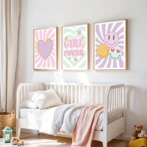 May include: Three framed wall art prints with a retro theme. The prints feature a heart, the words "GIRL POWER", and smiley faces. Each print has a light-colored background with a sunburst design. The frames are a light wood color.