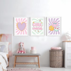 May include: Three framed wall art prints with white frames. The prints feature a heart, the words "GIRL POWER", and smiley faces. The artwork has a retro aesthetic with pastel colors and sunburst backgrounds. The prints are displayed above a bench with a stuffed unicorn.